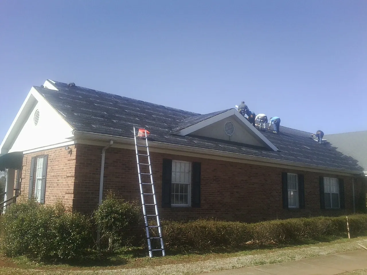 Crew tearing shingles off a brick ranch roof with a ladder set — Metal Roof Repair work by our crew in Houghton