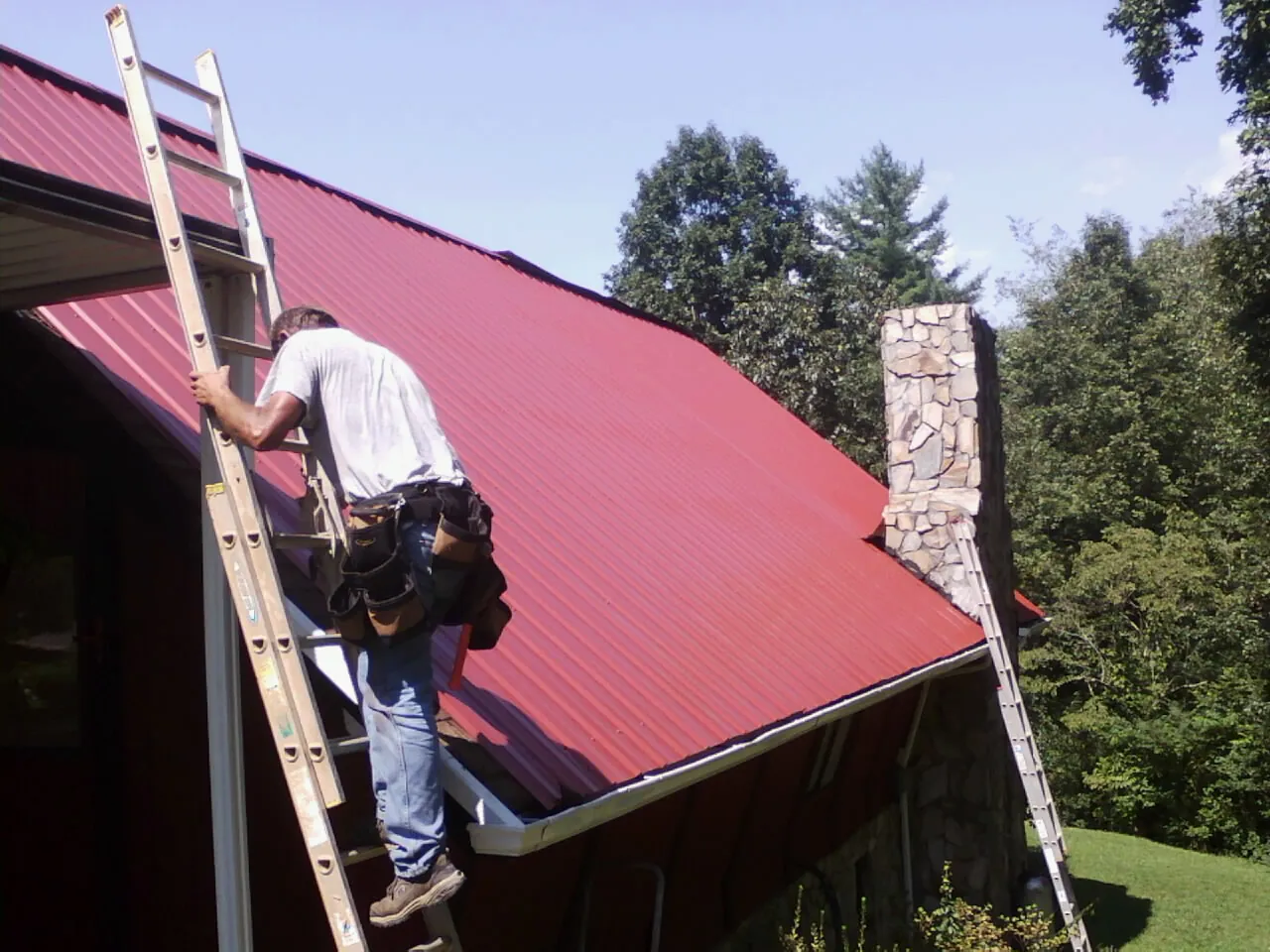 Roofer on a ladder beside a red metal roof and stone chimney — Soffit & Fascia Repair by Roofing Brothers in Houghton